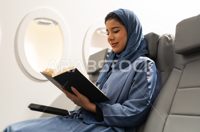 Enjoying reading books, stories, and novels, the culture of self-learning, practicing the hobby of reading, spending free time learning, an Arab Gulf Omani woman wearing a hijab and abaya sitting in a chair inside an airplane on a flight holding a book in her hand.