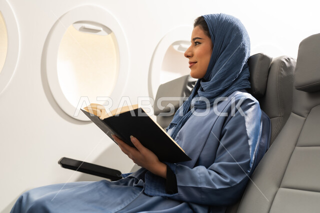 Spending free time learning, enjoying reading books, stories, and novels, the culture of self-learning, practicing the hobby of reading, an Arab Gulf Omani woman wearing a hijab and abaya sitting in a chair inside an airplane on a flight, holding a book in her hand and looking at something.