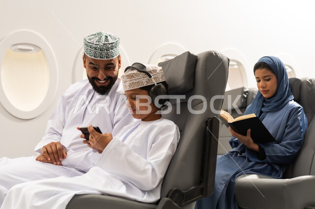 قضاء وقت الفراغ بالتعلم والثقافة, رحلة جوية سياحية , ولد عربي خليجي عماني يرتدي الدشداشة و الكمه يجلس على مقعده داخل الطائرة برفقه والده يمسك الهاتف المحمول , سيده خليجيه تقوم بقراءة كتاب بتعابير التركيز والتمعن