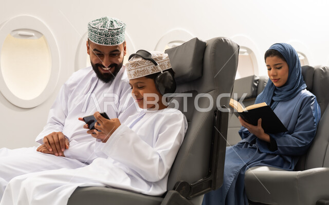 هواية المطالعة والاستكشاف, رحلة جوية سياحية , ولد عربي خليجي عماني يرتدي الدشداشة و الكمه يجلس على مقعده داخل الطائرة برفقه والده يمسك الهاتف المحمول , سيده خليجيه تقوم بقراءة كتاب بتعابير التركيز والتمعن