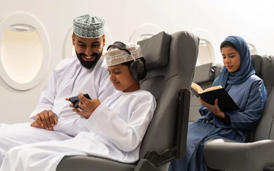 هواية المطالعة والاستكشاف, رحلة جوية سياحية , ولد عربي خليجي عماني يرتدي الدشداشة و الكمه يجلس على مقعده داخل الطائرة برفقه والده يمسك الهاتف المحمول , سيده خليجيه تقوم بقراءة كتاب بتعابير التركيز والتمعن