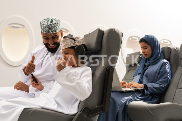 A tourist flight, the concept of acceptance and approval, an Omani Gulf Arab boy wearing a dishdasha and a kumma sits in his seat inside the plane with his father, holding a mobile phone with gestures of victory and triumph, a Gulf woman working on a laptop.