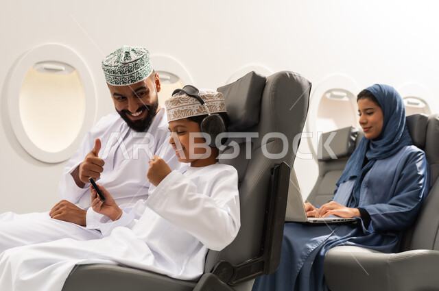 الذهاب في جولات ترفيهية في إجازة الصيف, مفهوم القبول والموافقه , ولد عربي خليجي عماني يرتدي الدشداشة و الكمه يجلس على مقعده داخل الطائرة برفقه والده يمسك الهاتف المحمول بايماءات الفوز و الانتصار , سيده خليجيه تقوم بالعمل على الحاسب المحمول