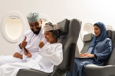 الذهاب في جولات ترفيهية في إجازة الصيف, مفهوم القبول والموافقه , ولد عربي خليجي عماني يرتدي الدشداشة و الكمه يجلس على مقعده داخل الطائرة برفقه والده يمسك الهاتف المحمول بايماءات الفوز و الانتصار , سيده خليجيه تقوم بالعمل على الحاسب المحمول