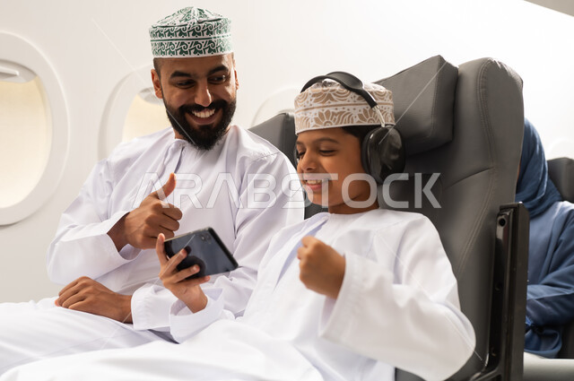 The concept of acceptance and agreement, going on recreational tours during the summer vacation, an Arab Gulf Omani boy wearing a dishdasha and a kumma sitting in his seat inside the airplane with his father holding a mobile phone with gestures of victory and triumph, Oman Air.