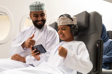 مفهوم القبول والموافقه, الذهاب في جولات ترفيهية في إجازة الصيف , ولد عربي خليجي عماني يرتدي الدشداشة و الكمه يجلس على مقعده داخل الطائرة برفقه والده يمسك الهاتف المحمول بايماءات الفوز و الانتصار , خطوط الطيران العمانية