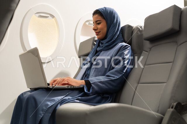 القيام بجولات ترفيهية في إجازة الصيف,الشعور بالسعادة والفرح, سيده عربيه خليجيه عمانية ترتدي الحجاب و العباءه تجلس على كرسي داخل الطائرة فى رحلة جويه تقوم باستخدام الحاسب المحمول, استخدام جهاز تقني حديث ومتطور