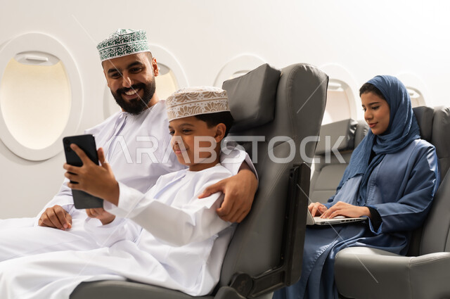 رحلة صيفية ترفيهية ممتعة, سيده عربيه خليجيه محجبه تقوم باستخدام الحاسب المحمول, ولد عربي خليجي يرتدي الدشداشة و الكمه يجلس داخل الطائرة برفقة والده يقوم بالتقاط صورة سيلفي عبر الهاتف المحمول , ايماءات السعاده و السرور