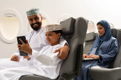 رحلة صيفية ترفيهية ممتعة, سيده عربيه خليجيه محجبه تقوم باستخدام الحاسب المحمول, ولد عربي خليجي يرتدي الدشداشة و الكمه يجلس داخل الطائرة برفقة والده يقوم بالتقاط صورة سيلفي عبر الهاتف المحمول , ايماءات السعاده و السرور