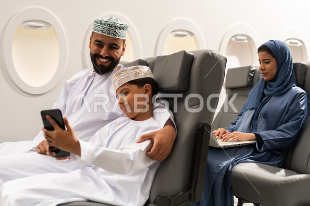 قضاء رحلة صيفية ممتعة, الاستعداد لبدء العطلة خارج البلاد,سيده عربيه خليجيه محجبه تقوم باستخدام الحاسب المحمول, ولد عربي خليجي يرتدي الدشداشة و الكمه يجلس داخل الطائرة برفقة والده يقوم بالتقاط صورة سيلفي عبر الهاتف المحمول , ايماءات السعاده و السرور