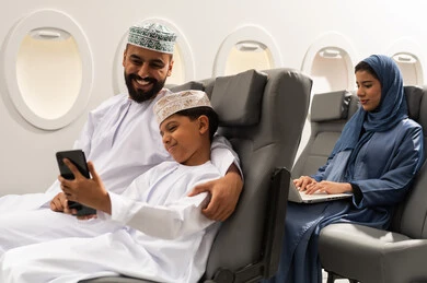 Spending an enjoyable summer trip, preparing to start the vacation abroad, a Gulf Arab woman in a hijab is using a laptop, a Gulf Arab boy wearing a dishdasha and a kuffiyeh is sitting inside the airplane with his father taking a selfie with a mobile phone, gestures of happiness and joy.