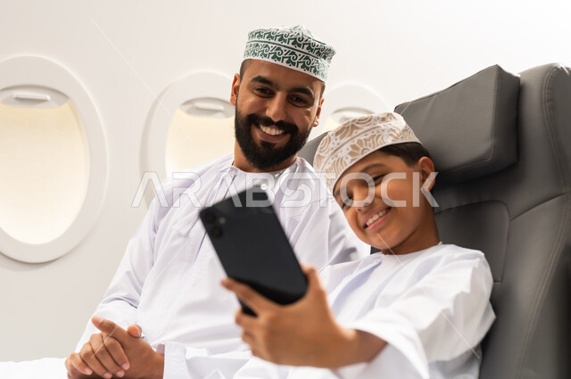 الاستعداد لبدء العطلة خارج البلاد, قضاء رحلة صيفية ممتعة, السياحة في إجازة الصيف, ولد عربي خليجي يرتدي الدشداشة و الكمه يجلس داخل الطائرة برفقة والده  يقوم بالتقاط صورة سيلفي عبر الهاتف المحمول , ايماءات السعاده و السرور