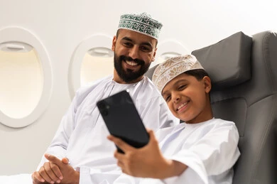 الاستعداد لبدء العطلة خارج البلاد, قضاء رحلة صيفية ممتعة, السياحة في إجازة الصيف, ولد عربي خليجي يرتدي الدشداشة و الكمه يجلس داخل الطائرة برفقة والده  يقوم بالتقاط صورة سيلفي عبر الهاتف المحمول , ايماءات السعاده و السرور