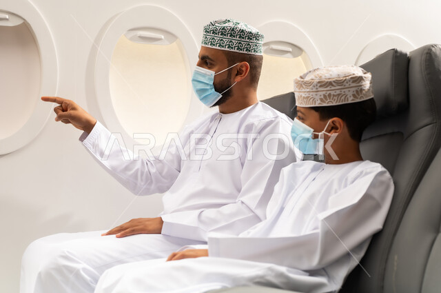 قضاء أوقات ممتعة داخل الطائرة, رجل عربي خليجي عماني يرتدي الدشداشة و الكمه و قناع الحماية من فايروس كورونا يجلس على مقعده فى رحله جويه برفقه ابنه و يقوم بالاشارة الى شي ما , عطلة صيفية عائلية، الخطوط الجوية العمانيه، سياحة وسفر