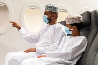 قضاء أوقات ممتعة داخل الطائرة, رجل عربي خليجي عماني يرتدي الدشداشة و الكمه و قناع الحماية من فايروس كورونا يجلس على مقعده فى رحله جويه برفقه ابنه و يقوم بالاشارة الى شي ما , عطلة صيفية عائلية، الخطوط الجوية العمانيه، سياحة وسفر