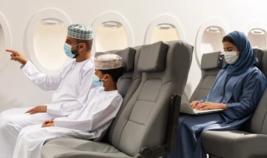 الاشارة الى شيء ما , عائله عربيه خليجيه عمانية ترتدي الزي التقليدي  و قناع الحمايه ضد فايروس كورونا تجلس داخل الطائرة فى رحله جوية , امراة خليجيه محجبه تقوم بالعمل على الحاسب المحمول , مفهوم السياحة و السفر
