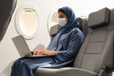Performing tasks and work duties, integrating and focusing to accomplish tasks remotely, a close-up side view of an Arab Gulf Omani woman wearing an abaya and hijab and a coronavirus mask sitting in her seat inside the airplane on a flight working on a laptop.