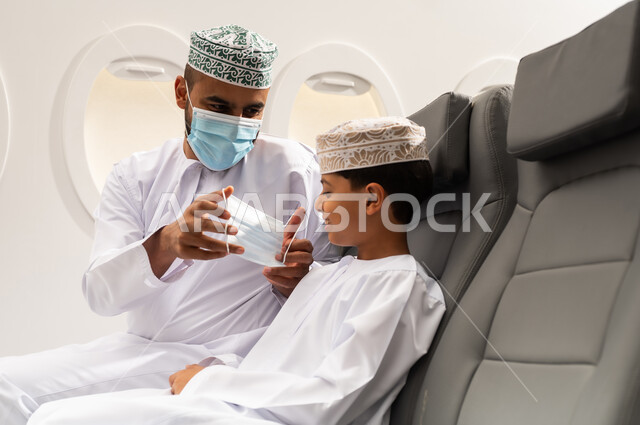 اتباع الإجراءات الاحترازية الوقائية من فايروس كورونا، رجل عماني خليجي يرتدي الدشداشة والكمة يجلس على مقعده في الطائرة برفقة ابنه، ولد خليجي يرتدي قناع الحماية، رحلة عائلية، مفهوم السياحة والسفر