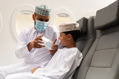 اتباع الإجراءات الاحترازية الوقائية من فايروس كورونا، رجل عماني خليجي يرتدي الدشداشة والكمة يجلس على مقعده في الطائرة برفقة ابنه، ولد خليجي يرتدي قناع الحماية، رحلة عائلية، مفهوم السياحة والسفر