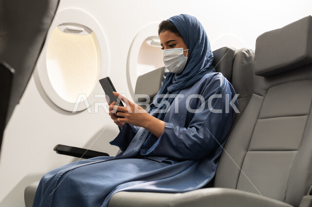 استخدام جهاز تقني حديث ومتطور, تبادل الأحاديث والأخبار مع الأهل والأصدقاء، امراة  عربيه خليجيه عمانية ترتدي العباءة والحجاب و ماسك كورونا تمسك بيدها الهاتف المحمول ، قضاء وقت ممتع داخل الطائرة، تصفح التطبيقات وبرامج التواصل الاجتماعي