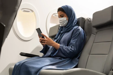 استخدام جهاز تقني حديث ومتطور, تبادل الأحاديث والأخبار مع الأهل والأصدقاء، امراة  عربيه خليجيه عمانية ترتدي العباءة والحجاب و ماسك كورونا تمسك بيدها الهاتف المحمول ، قضاء وقت ممتع داخل الطائرة، تصفح التطبيقات وبرامج التواصل الاجتماعي
