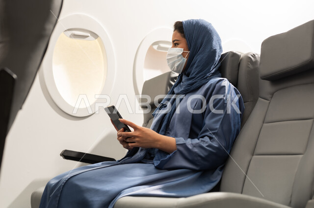 تبادل الأحاديث والأخبار مع الأهل والأصدقاء، امراة  عربيه خليجيه عمانية ترتدي العباءة والحجاب و قناع كورونا تمسك بيدها الهاتف المحمول ، استخدام جهاز تقني حديث ومتطور، قضاء وقت ممتع داخل الطائرة، تصفح التطبيقات وبرامج التواصل الاجتماعي