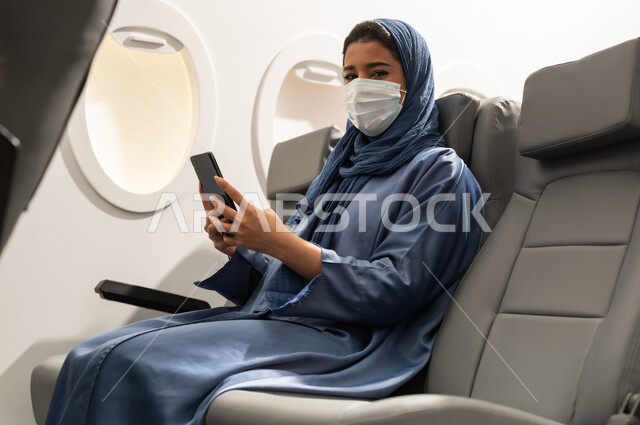 Wearing a medical mask to prevent the coronavirus, airlines, tourism and travel, an Arab Gulf Omani woman wearing an abaya and hijab sitting in her seat inside the airplane holding a mobile phone.