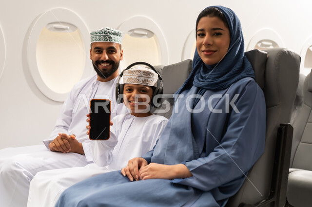 عرض شاشة فارغه سوداء بالهاتف المحمول , دمج التقنيات فى الحياه اليوميه ،الترفيه والتسليه وقضاء الاوقات داخل الطائره ، عائله عربيه خليجيه عمانية ترتدي الزي التقليدي تجلس على كراسي الطائره في رحله جويه