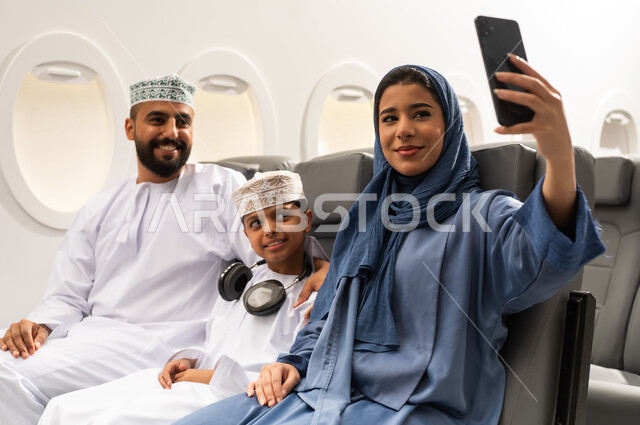 التقاط صور السيلفي عبر الهاتف المحمول, دمج التقنيات فى الحياه اليوميه ،الترفيه والتسليه وقضاء الاوقات داخل الطائره  ، عائله عربيه خليجيه عمانية ترتدي الزي التقليدي تجلس على كراسي الطائره في رحله جويه