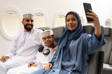 Taking selfies with a mobile phone, integrating technology into daily life, entertainment and leisure, and spending time inside the plane, an Omani Gulf Arab family wearing traditional attire sitting on the plane seats during a flight.