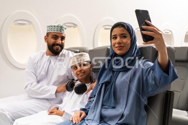 الترفيه والتسليه وقضاء الاوقات داخل الطائره, دمج التقنيات فى الحياه اليوميه  ، التقاط صور السيلفي عبر الهاتف المحمول ، عائله عربيه خليجيه عمانية ترتدي الزي التقليدي تجلس على كراسي الطائره في رحله جويه