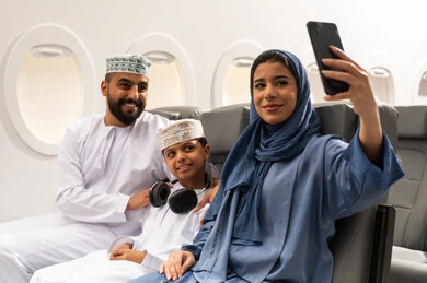 Entertainment and leisure and spending time inside the plane, integrating technology into daily life, taking selfies with a mobile phone, an Arab Gulf Omani family wearing traditional attire sitting on the plane seats during a flight.