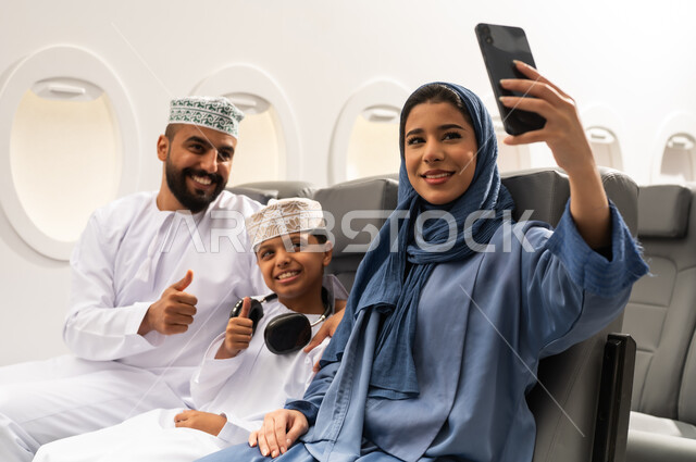 Integrating technology into daily life, entertainment and leisure, and spending time inside the plane, taking selfies with a mobile phone, an Omani Gulf Arab family wearing traditional attire sitting on the plane seats during a flight.
