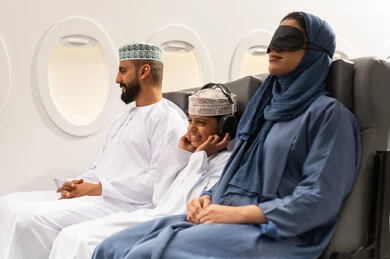 An Omani Gulf Arab family wearing traditional attire sits inside the airplane, showcasing hospitality, traveling with family, airline services, tourism and travel. A Gulf woman wearing a black eye mask is sleeping in her seat, displaying gestures of happiness and joy.