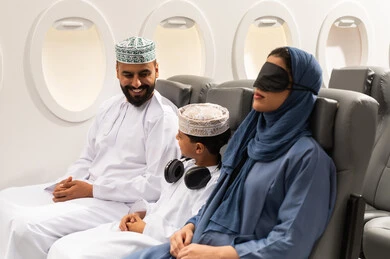 Tourist trip, airline lines, an Omani Gulf Arab family wearing traditional attire sitting inside the airplane, hospitality, tourism and travel, a Gulf woman wearing a black eye mask sleeping in her seat, gestures of happiness and joy.