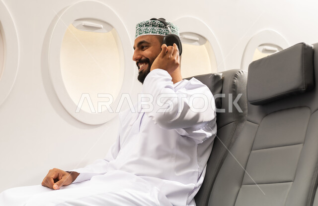التفاعل والاندماج مع الأنغام الموسيقية, الاستماع للأغاني باسخدام الهيدفون، شاب عربي خليجي عماني يرتدي الدشداشة والكمه يجلس على كرسي الطائرة فى رحلة جوية يضع سماعات الرأس بإيماءات الفرح والسرور