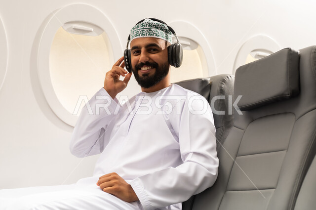 شاب عربي خليجي عماني يرتدي الدشداشة والكمه يجلس على كرسي الطائرة فى رحلة جوية يضع سماعات الرأس بإيماءات الفرح والسرور, الاستماع للأغاني باسخدام الهيدفون، التفاعل والاندماج مع الأنغام الموسيقية
