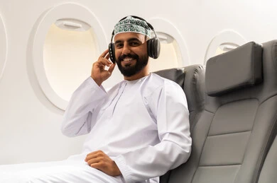 A young Arab Gulf Omani man wearing a dishdasha and kumma sits in an airplane seat on a flight, wearing headphones with gestures of joy and happiness, listening to music using the headphones, interacting and immersing himself in the musical tunes.