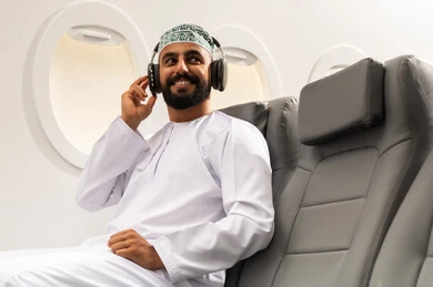 الاستماع للأغاني باسخدام الهيدفون، شاب عربي خليجي عماني يرتدي الدشداشة والكمه يجلس على كرسي الطائرة فى رحلة جوية يضع سماعات الرأس بإيماءات الفرح والسرور، التفاعل والاندماج مع الأنغام الموسيقية