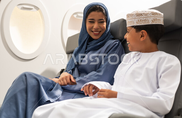 قضاء وقت ممتع ,التسليه والترفيه خلال ركوب الطائره, ولد عربي خليجي عماني يرتدي الدشداشة و الكمه يجلس برفقه والدته , الاستعداد لقضاء عطله صيفيه ممتعه , ايماءات السعادة و السرور