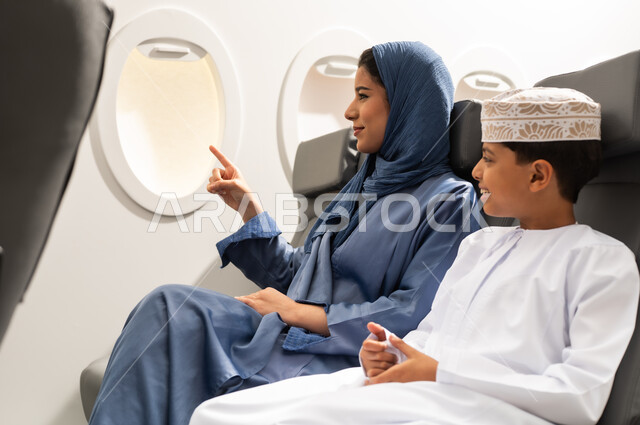 الاشارة الي شي ما ,التسليه والترفيه وقضاء الاوقات السعيده خلال ركوب الطائره, ولد عربي خليجي عماني يرتدي الدشداشة و الكمه يجلس برفقه والدته , الاستعداد لقضاء عطله صيفيه ممتعه , ايماءات السعادة و السرور