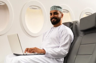 Training through the internet, using a laptop and working remotely, a side view of an Arab Gulf Omani man wearing a dishdasha and kumma sitting in a seat on a flight working on a laptop, the development of technology and technical devices, gestures of happiness and joy.