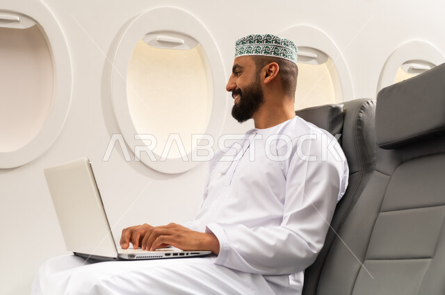 استخدام اللابتوب والعمل عن بعد, صورة من الجانب لرجل عربي خليجي عماني يرتدي الدشداشة و الكمه يجلس على المقعد فى رحله جويه يقوم بالعمل على الحاسب المحمول , تطور التكنولوجيا والأجهزة التقنية, التدريب من خلال الإنترنت, ايماءات السعاده و السرور