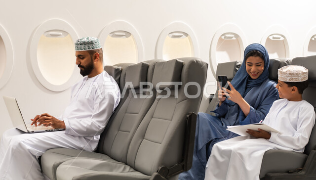استخدام التقنية الحديثة فى الحياه اليومية , عائله عربيه خليجيه عمانيه ترتدي الزي التقليدي تجلس داخل الطائرة فى رحله جويه , رجل خليجي يمسك بيده الحاسب المحمول , ام خليجيه تقوم بتصفح الهاتف المحمول بجانب ابنها , ايماءات السعادة و السرور