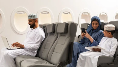 استخدام التقنية الحديثة فى الحياه اليومية , عائله عربيه خليجيه عمانيه ترتدي الزي التقليدي تجلس داخل الطائرة فى رحله جويه , رجل خليجي يمسك بيده الحاسب المحمول , ام خليجيه تقوم بتصفح الهاتف المحمول بجانب ابنها , ايماءات السعادة و السرور