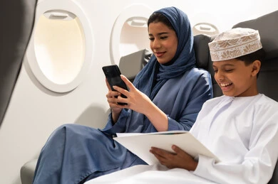قضاء أوقات ممتعة داخل الطائرة, امراة عربيه خليجيه عمانية ترتدي الحجاب و العباءه تجلس على المقعد فى رحله جويه تمسك بيدها الهاتف المحمول , ولد خليجي يجلس بجانب والدته و يقوم بالتلوين و الرسم , ايماءات السعادة و السرور
