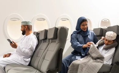 The concept of sleep and rest in the tourist journey, an Omani Gulf Arab family wearing traditional attire sitting in their seats on a flight, a Gulf Arab mother covering her child, a Gulf man holding a mobile phone, spending the summer vacation traveling and tourism, Oman Air.