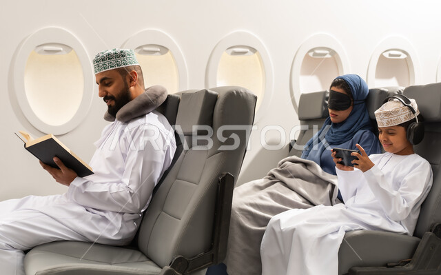 التسليه والترفيه وقضاء الاوقات السعيده خلال ركوب الطائره, عائله عربيه خليجيه عمانيه ترتدي الزي التقليدي يجلسون على مقاعدهم فى رحله جويه, ولد خليجي يقوم بالعب على الهاتف المحمول بجانب والدته , رجل عربى خليجي عماني يقوم بقراءة كتاب بايماءات التركيز