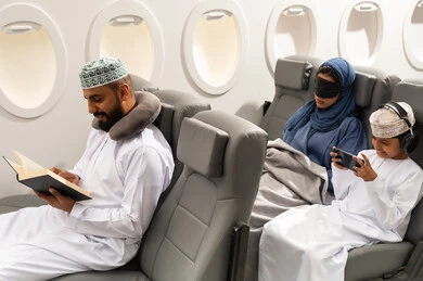 The concept of travel and tourism, an Arab Gulf Omani family dressed in traditional attire sitting in their seats inside one of the Gulf airlines, a Gulf boy playing on a mobile phone next to his mother, an Arab Gulf Omani man reading a book with gestures of concentration.