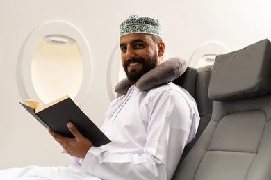 ايماءات السعادة و السرور , تنمية وزيادة الذخيرة الفكرية، الاهتمام بتصفح الكتب المفيدة، تمضية أوقات الفراغ بالتعلم والتثقيف، حب المطالعة والقراءة، رجل عربي خليجي عماني يرتدي الدشداشه والكمه و مخده السفر يجلس على كرسي الطائرة فى رحله جويه يحمل بيده كتاب